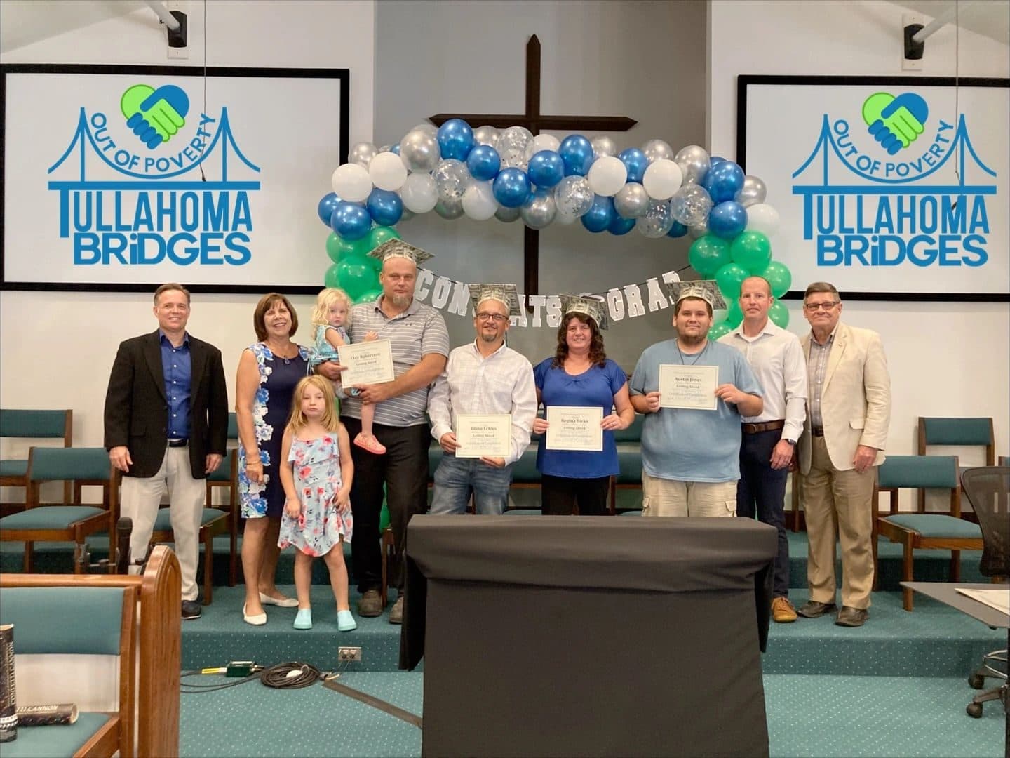 Getting Ahead graduates and their families on stage at the Tullahoma Bridges graduation ceremony, holding certificates under a balloon arch.